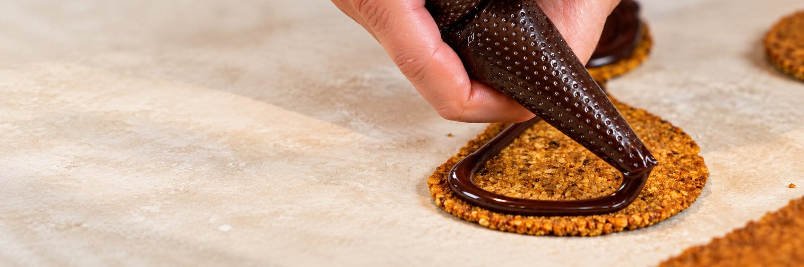 A hand piping melted chocolate onto a nut cookie on a floured countertop